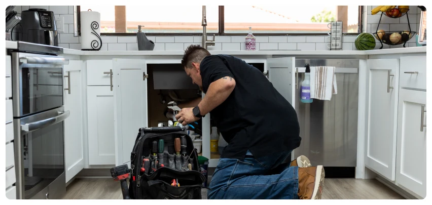 AC Installation 31 Technician kneeling under a kitchen sink, using tools for plumbing repair.